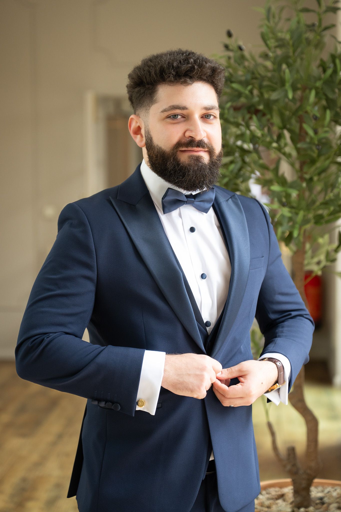 A person in a navy blue tuxedo and bow tie adjusts their suit jacket while getting ready for a formal event.
