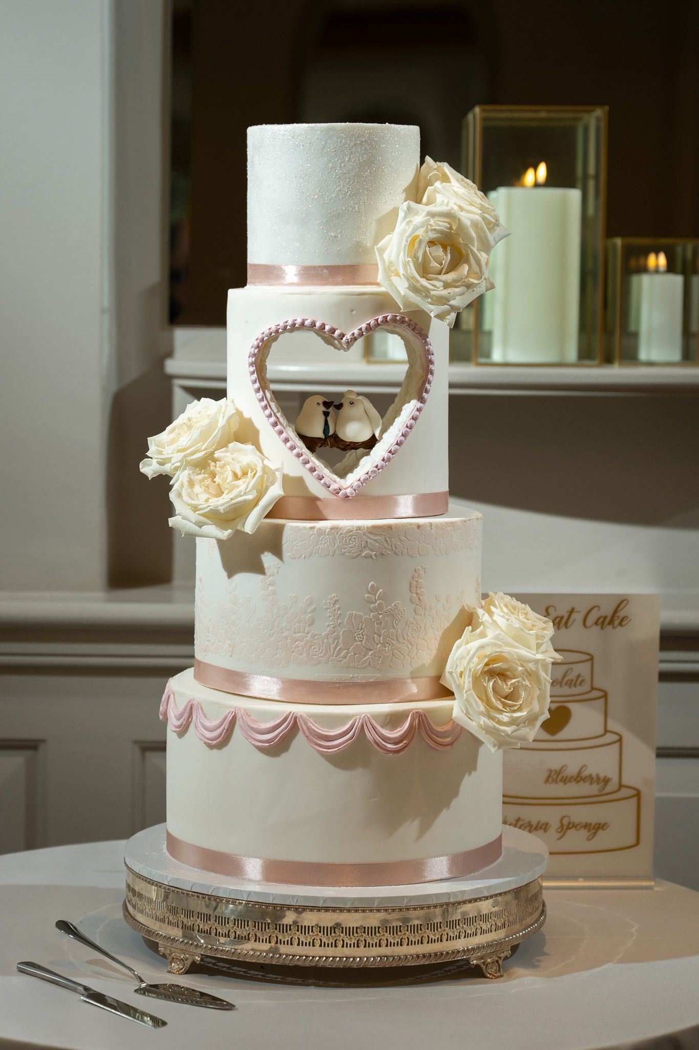 Elegant four-tier wedding cake with blush pink ribbons, heart detail, and cream roses on a silver cake stand.