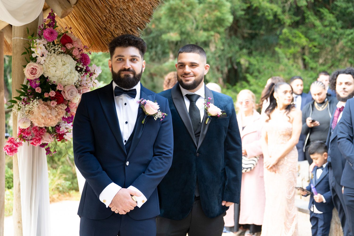 Wedding party members in navy suits and pink boutonnieres gather for formal photos in an outdoor garden setting.