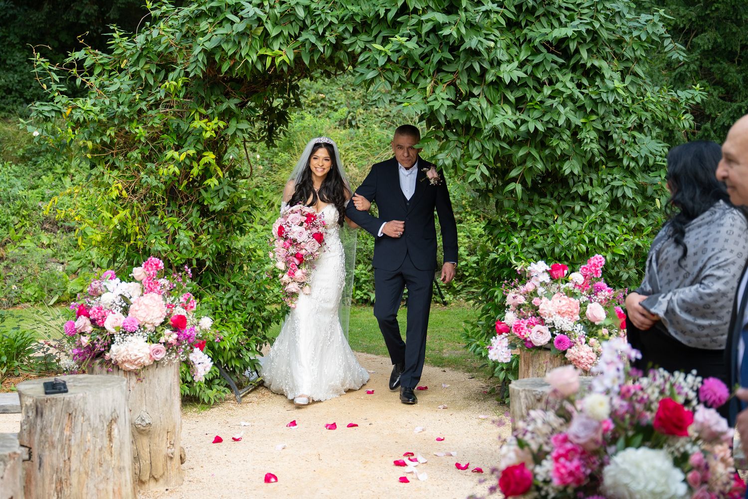 A couple walks along a garden path surrounded by lush greenery and pink floral arrangements during a celebration.
