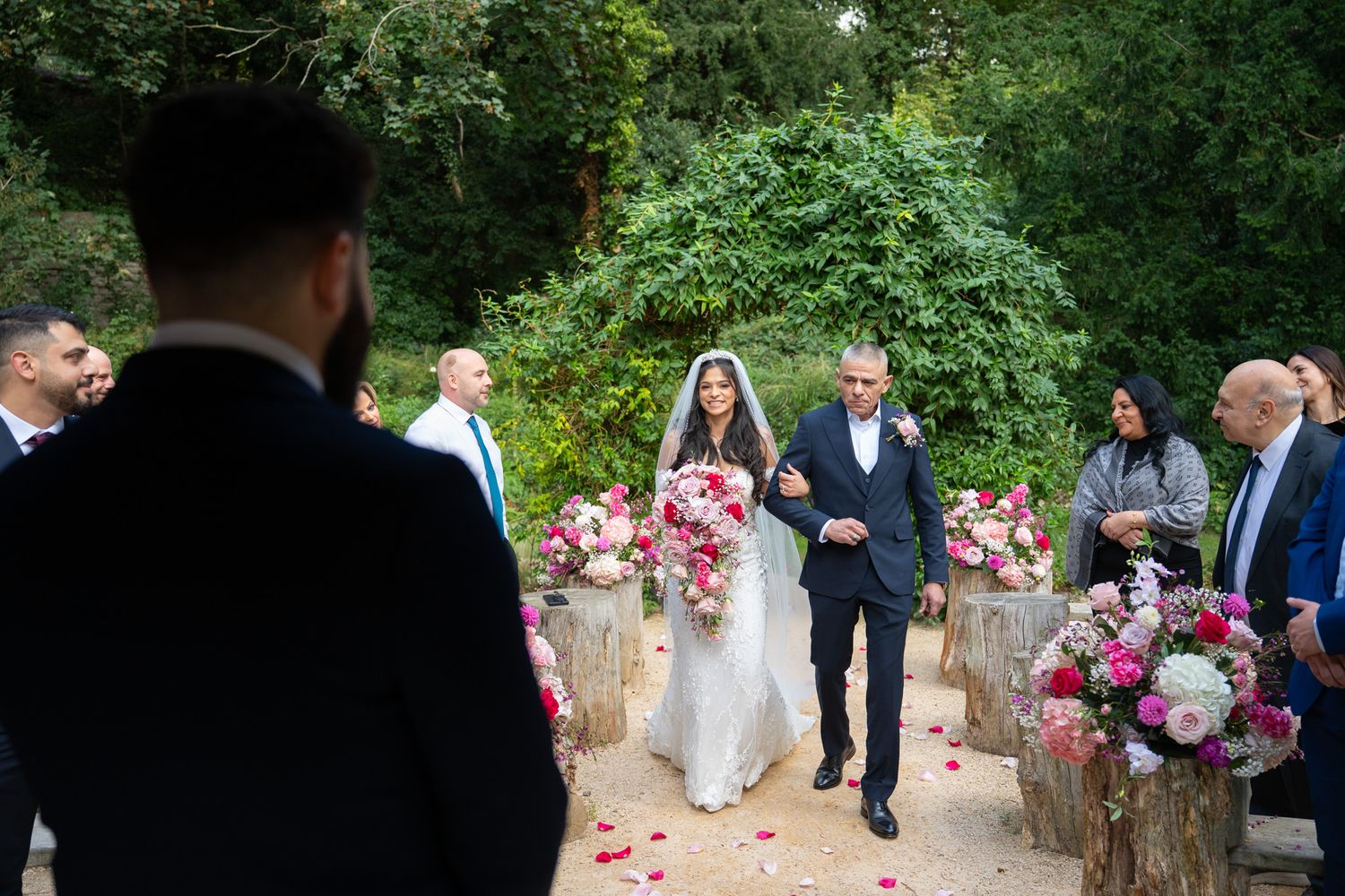 Outdoor wedding ceremony in a garden setting with pink floral arrangements on wooden stumps lining the aisle.