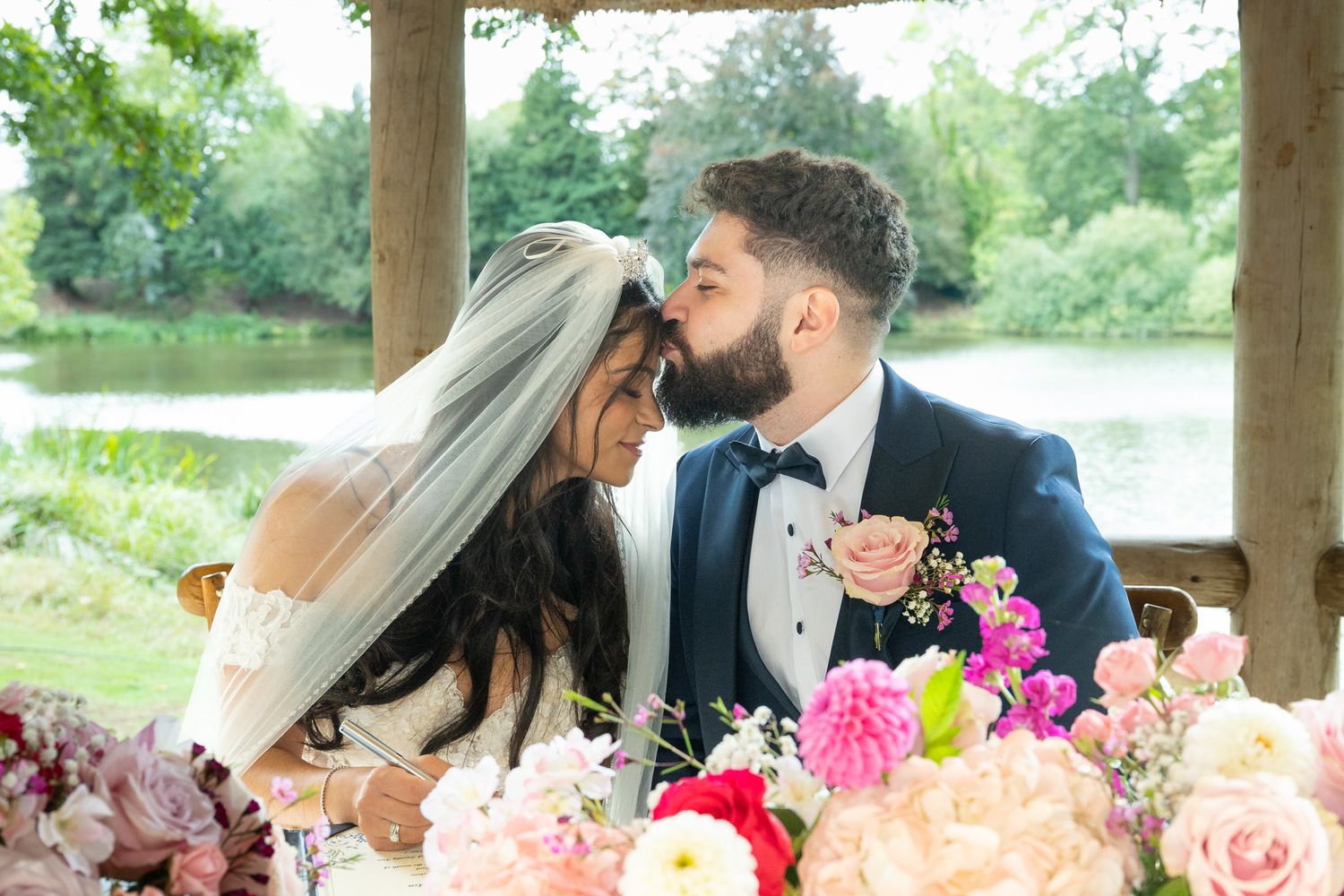 A romantic wedding couple shares tender moments under a rustic wooden pavilion surrounded by lush greenery and flowers.
