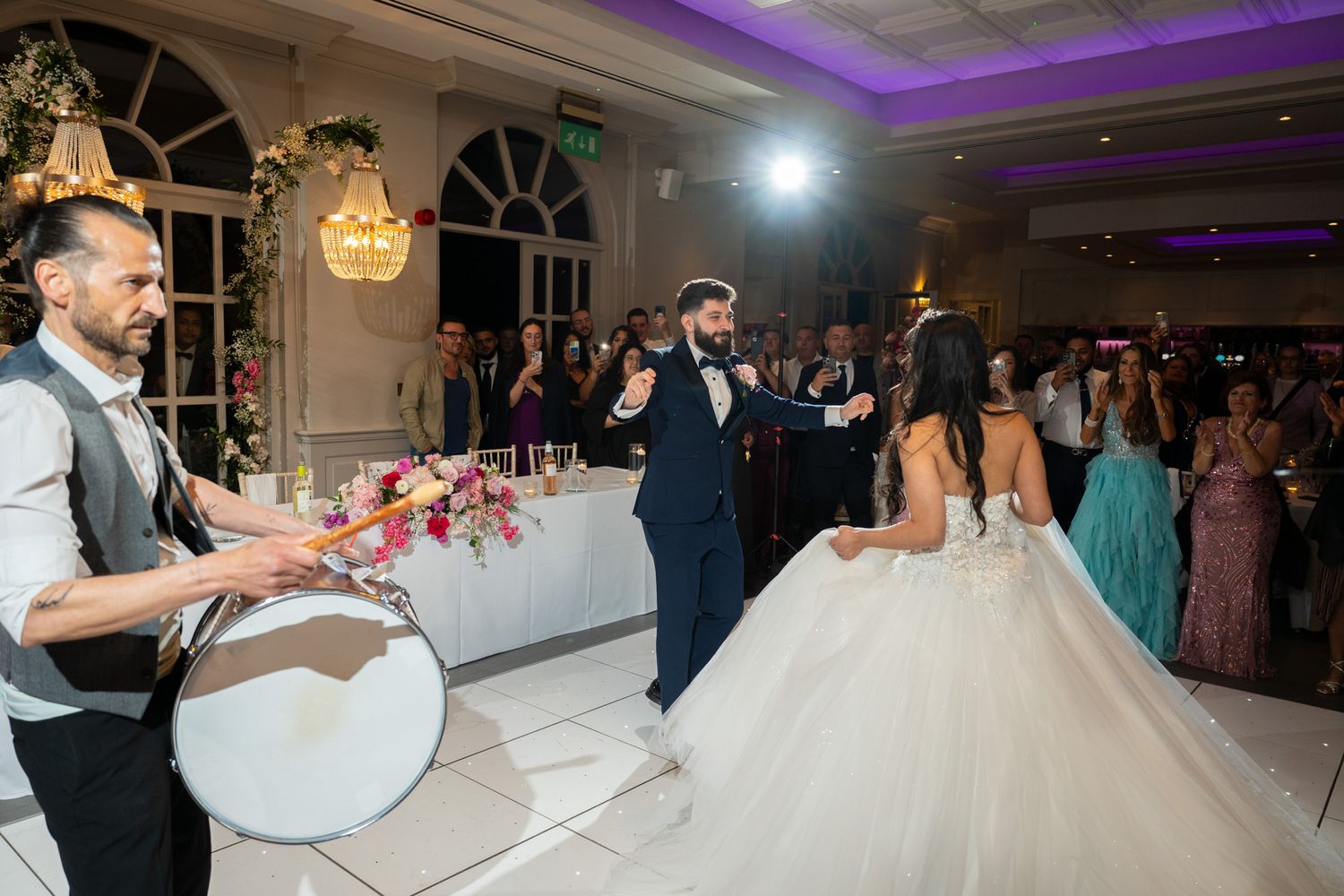 Wedding guests gather on the dance floor during a reception with purple uplighting in an elegant ballroom.
