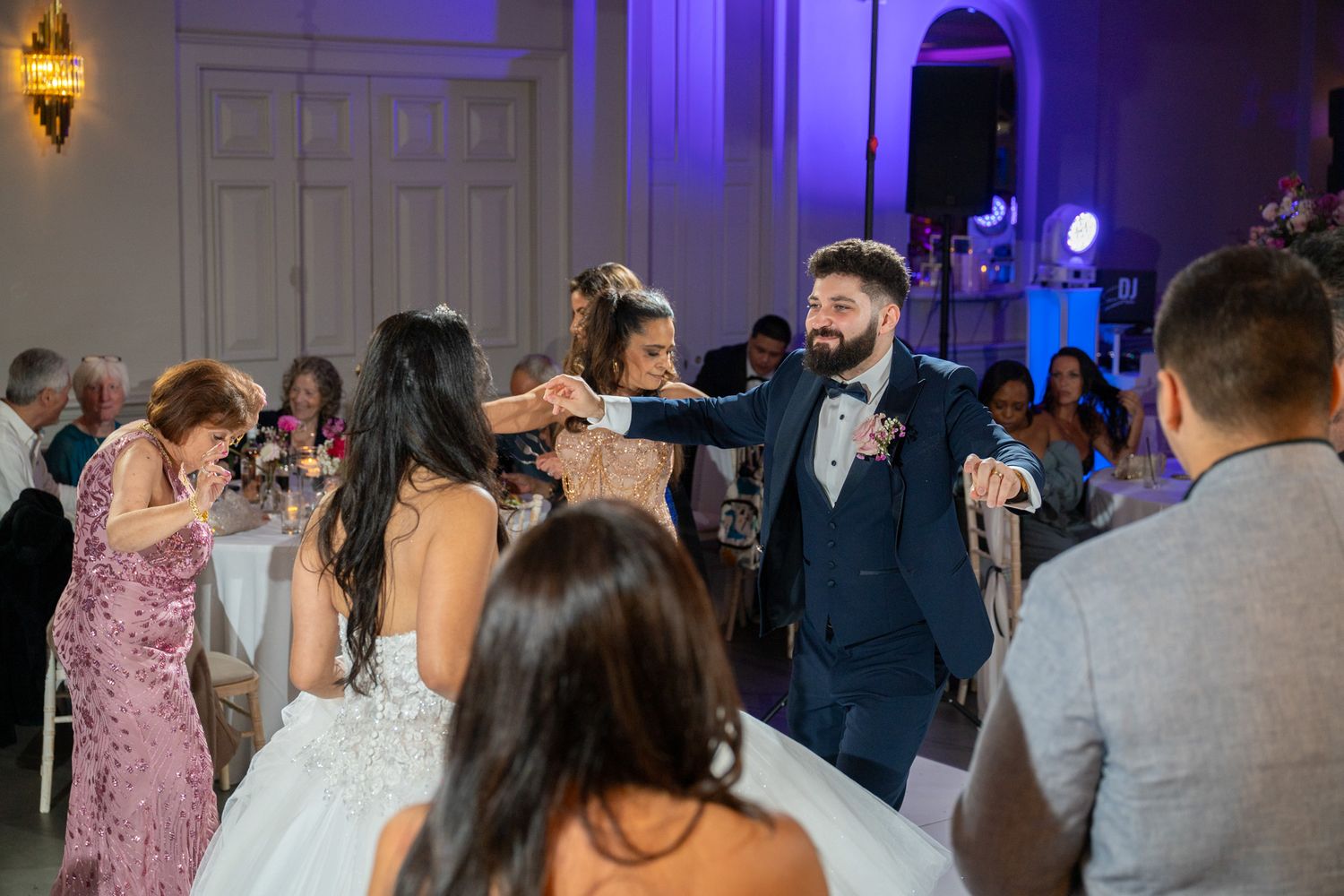 Wedding guests dance and celebrate on a dance floor illuminated by blue lighting in an elegant ballroom setting.