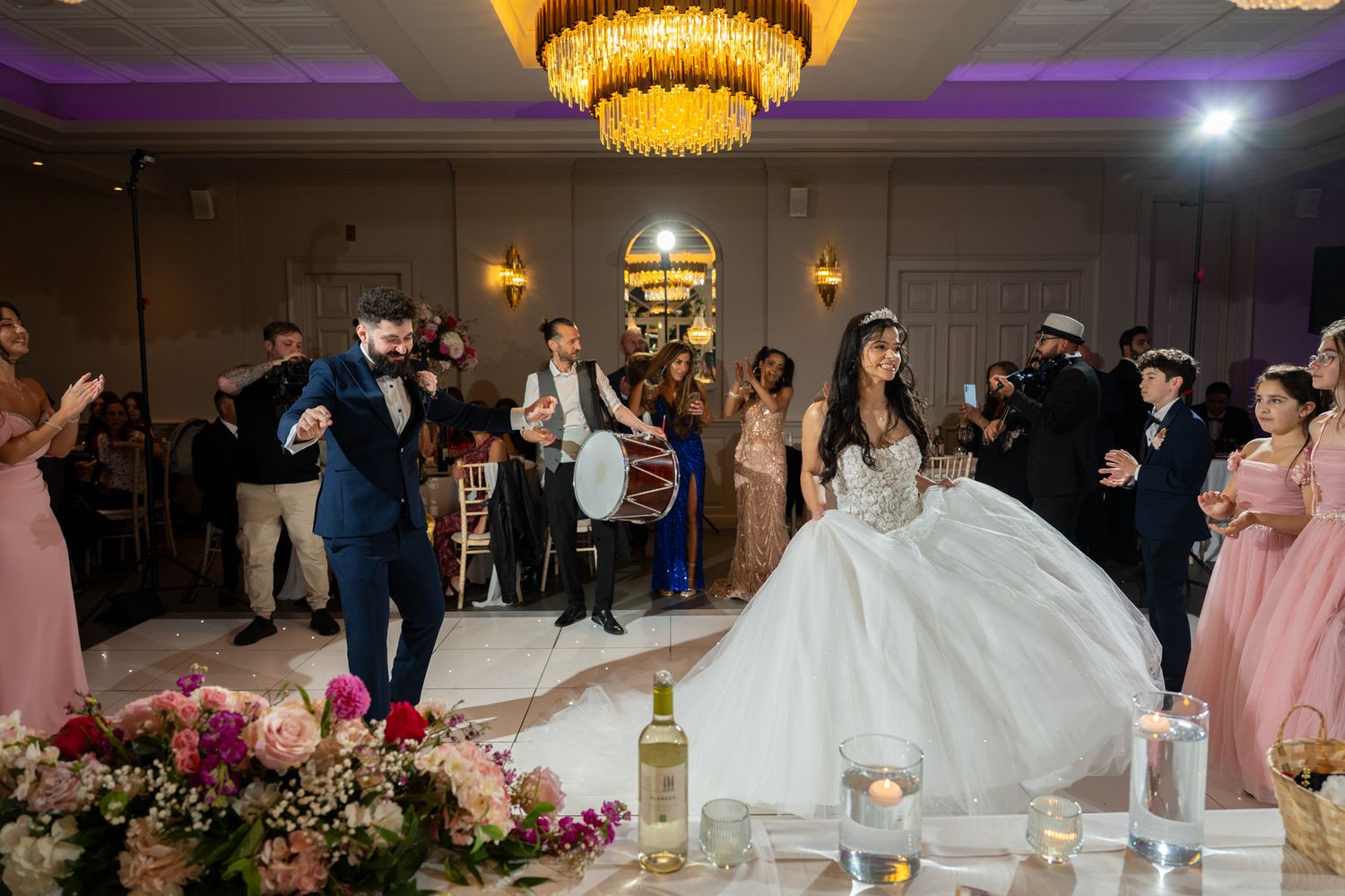 Wedding reception celebration in an elegant ballroom with guests dancing near floral centerpieces and chandeliers.