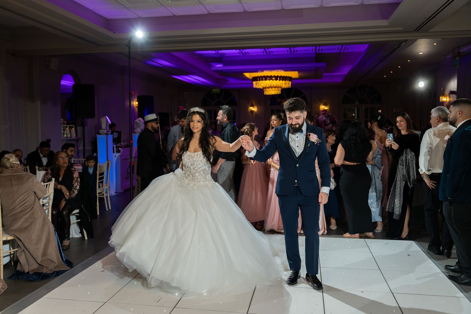 Wedding guests dance together on a purple-lit dance floor during an evening reception celebration.