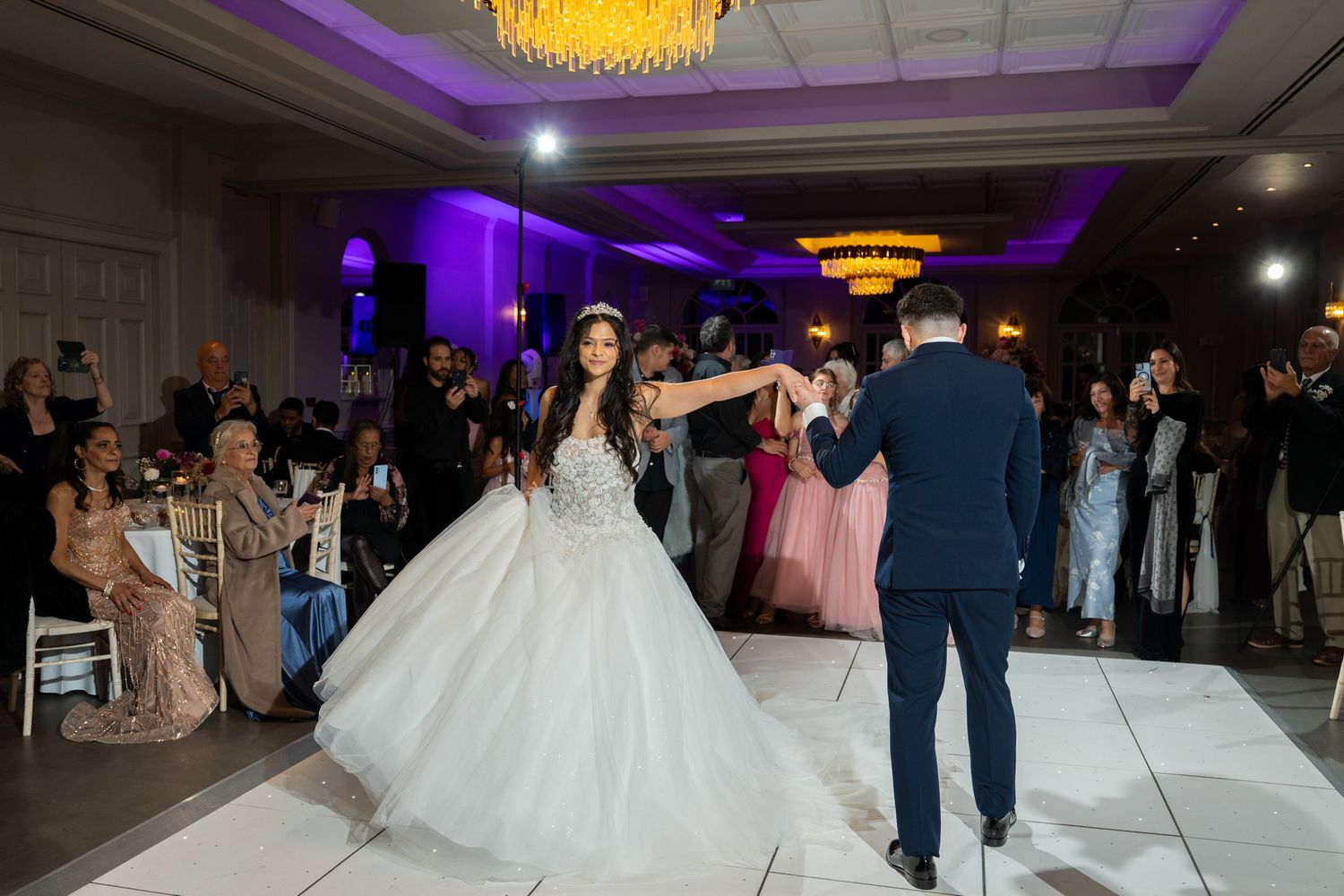 Wedding guests dancing together on an illuminated dance floor at an elegant reception venue with purple lighting.