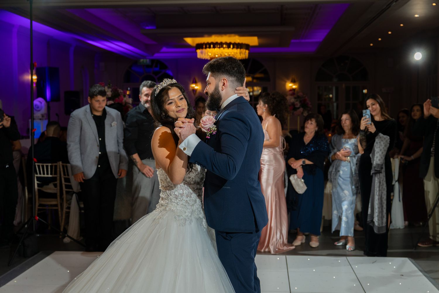 A couple shares their first dance at a wedding reception with purple lighting and guests watching from the sidelines.