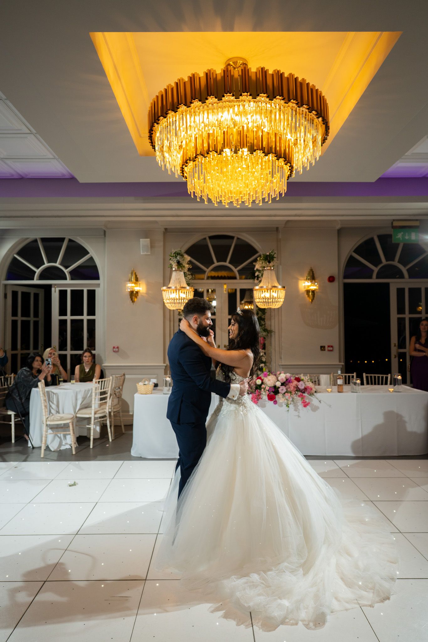 A couple dances elegantly beneath a crystal chandelier in an elegant ballroom with white decor and arched windows.
