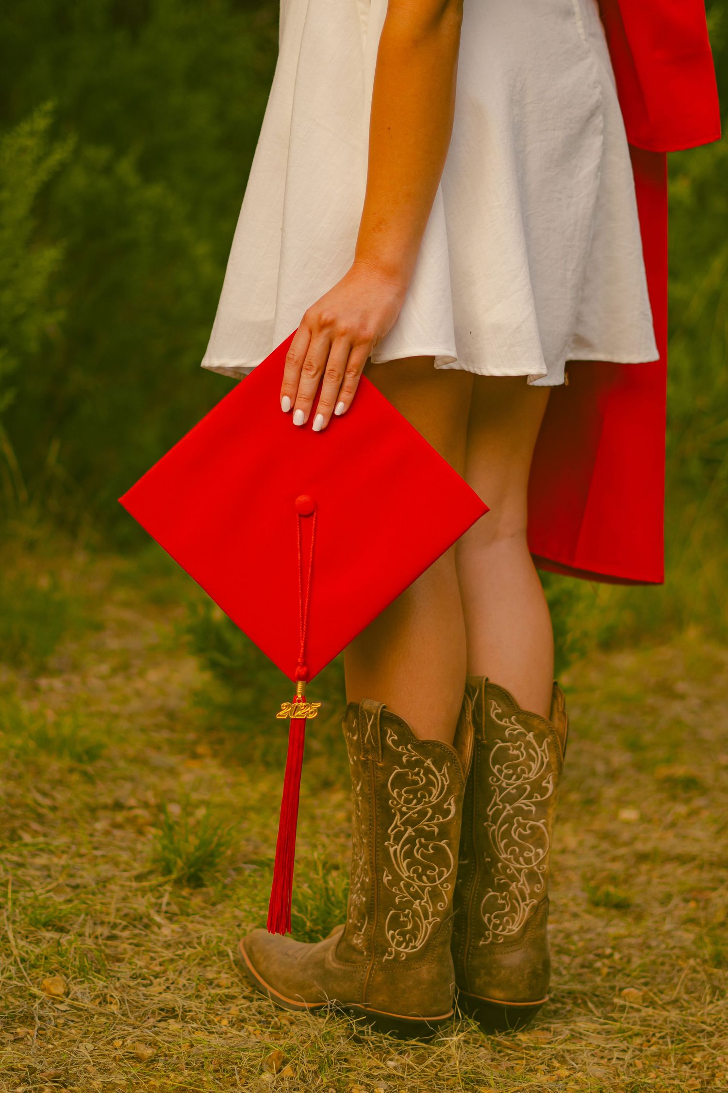 A graduate holds a red graduation cap while wearing decorative cowboy boots and a white dress.