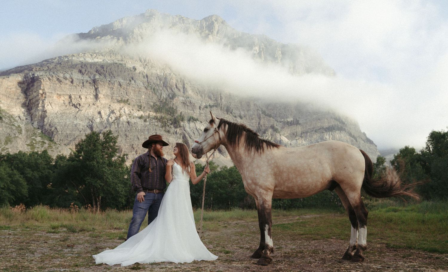 Bride in long train dress stands next to groom and buckskin horse while eloping in Glacier National Park.