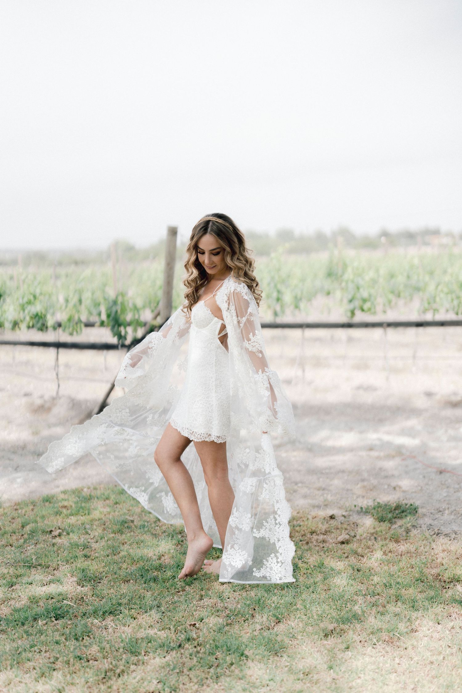 A bride in a flowing white dress adjusts her shoe while standing in a grassy outdoor setting.