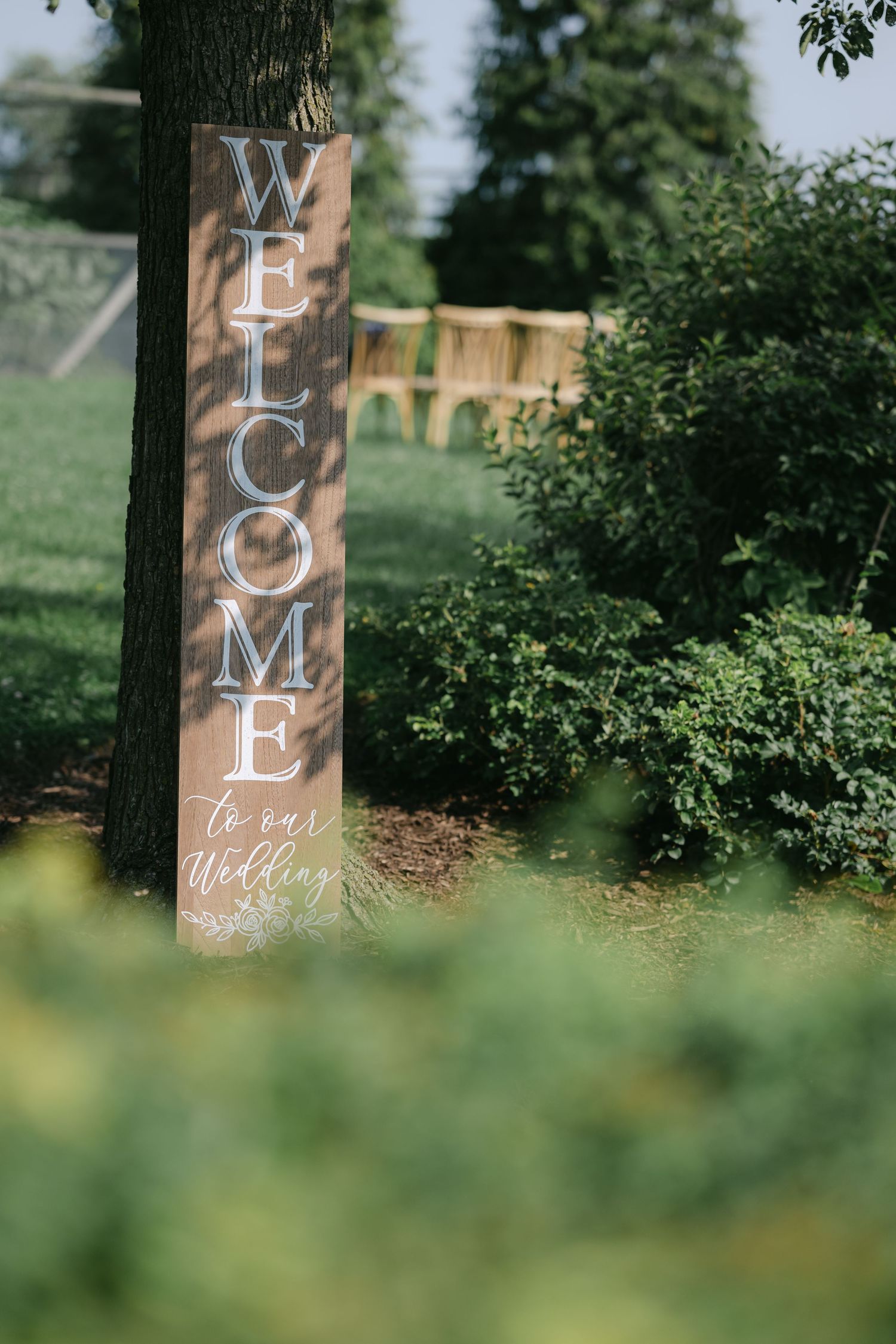 Rustic wooden welcome sign panels with white lettering displayed in a garden setting with green foliage in the background. - Elawa Farm