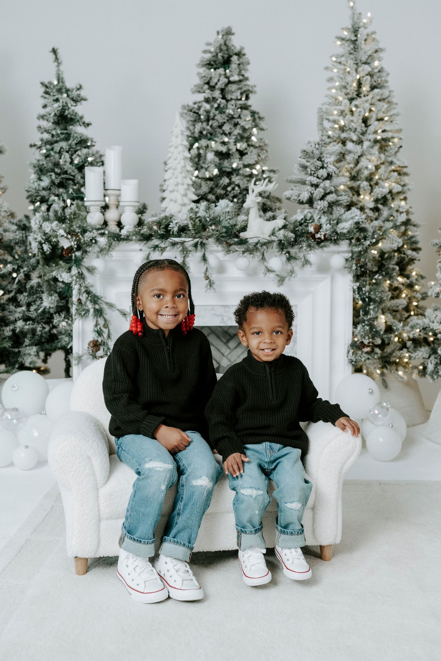 Two children sit on a white couch in front of flocked Christmas trees and white candle decorations in a winter scene.