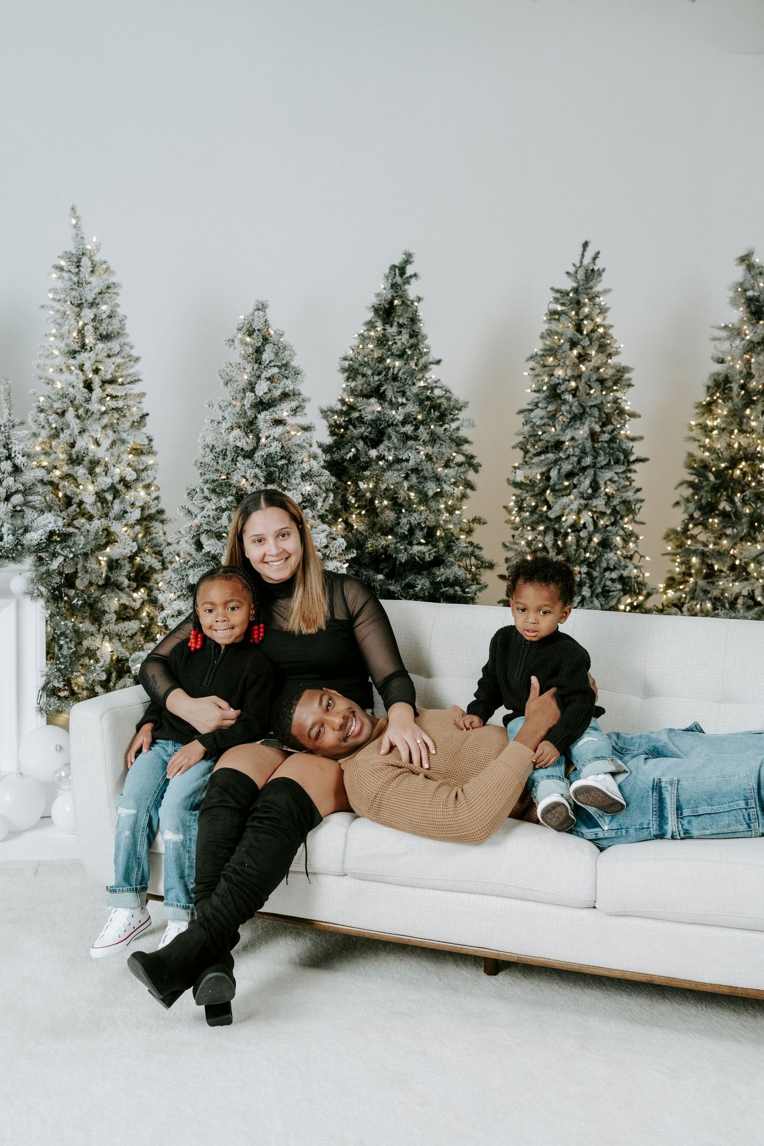A family lounges together on a white couch with decorated Christmas trees in the background.