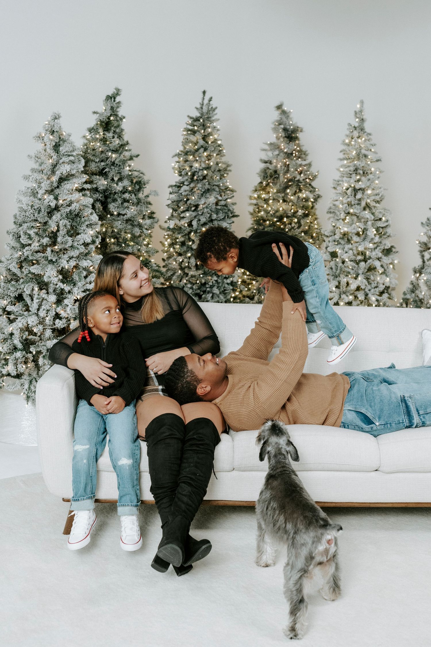 A couple lounges on a white couch with their dog while snow-covered evergreen trees are visible in the background.
