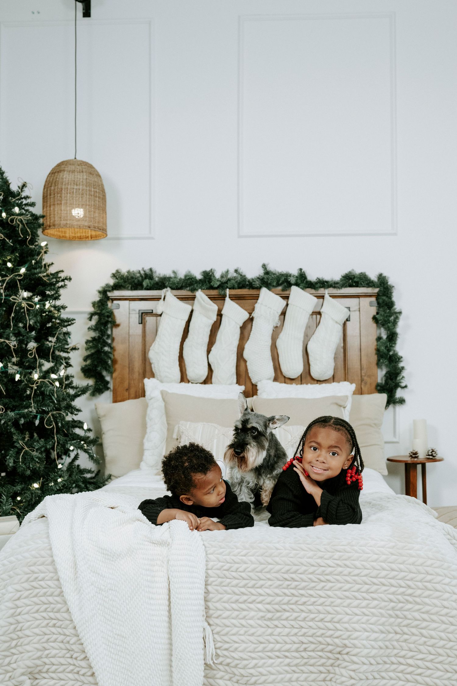 Three people snuggle under a chunky knit white blanket with Christmas stockings hanging on the wall behind them.