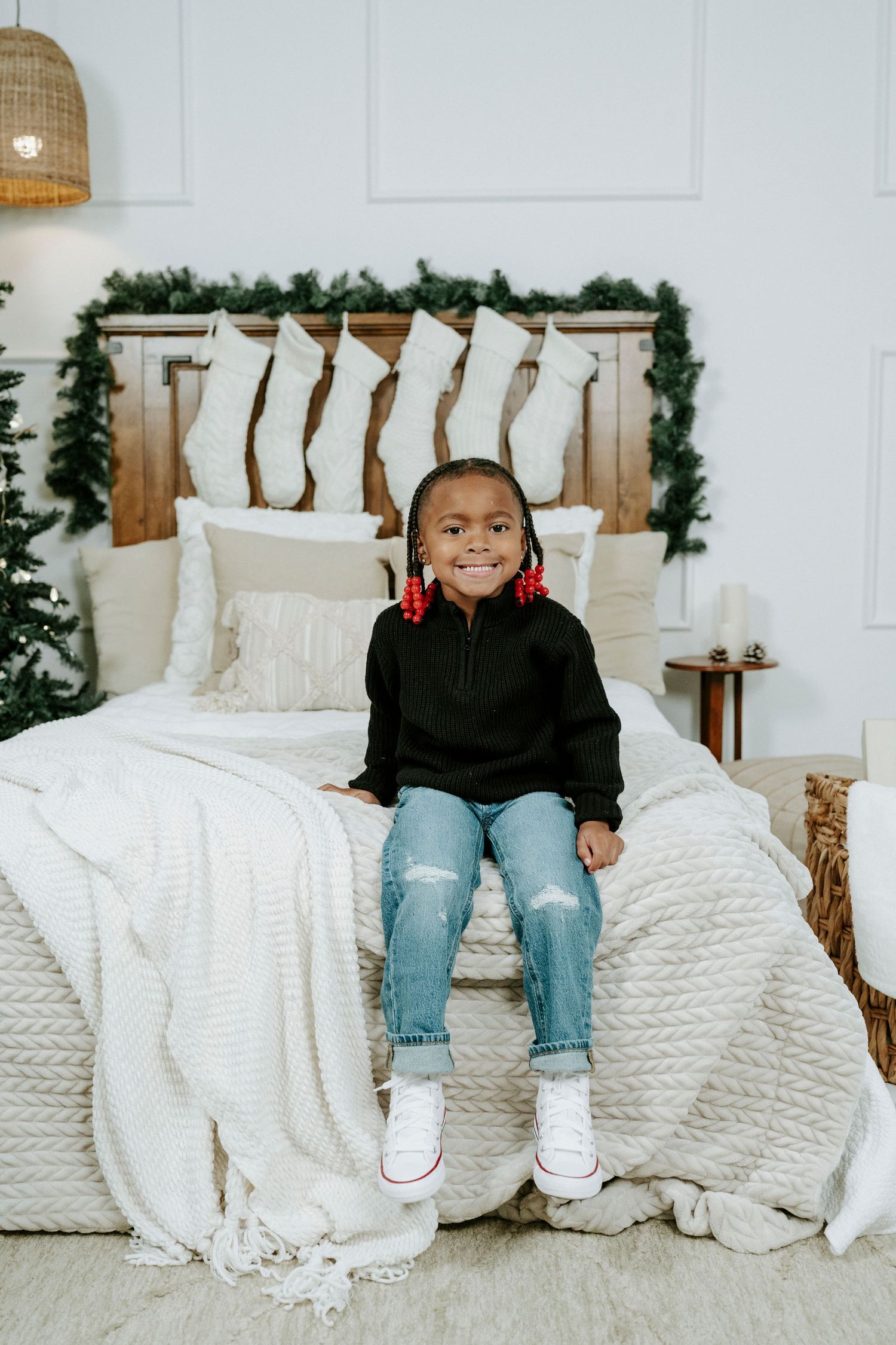 Child sits on white bedding with holiday greenery decor in a bright modern bedroom setting.