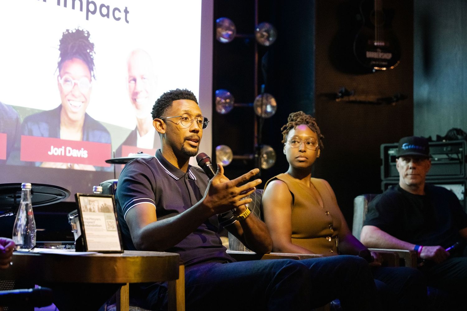 Three people sit at a panel discussion table with colorful lighting and bubbles floating in the dark background.