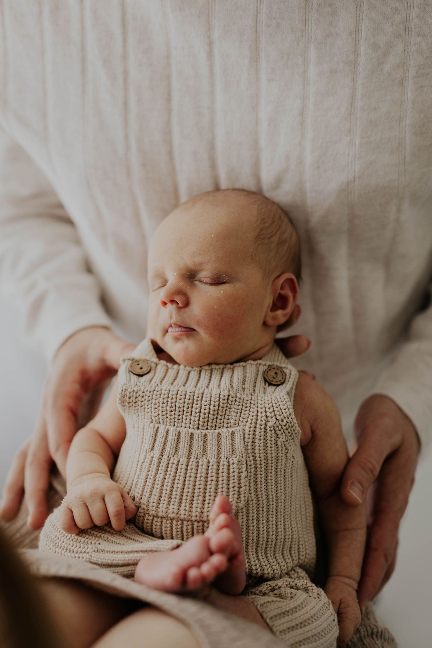 A newborn baby wearing a knit beige romper lies peacefully while gentle hands cradle him during newborn session in Lakeville MN