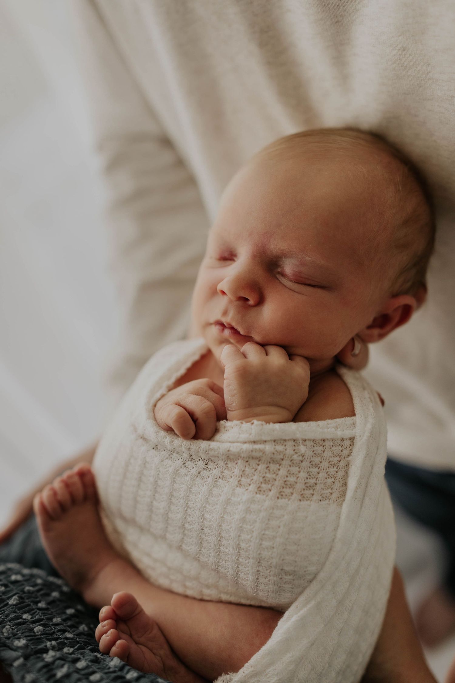 A sleeping newborn baby swaddled in white knit blanket lies peacefully in parents arms during newborn session near St. Paul, MN.
