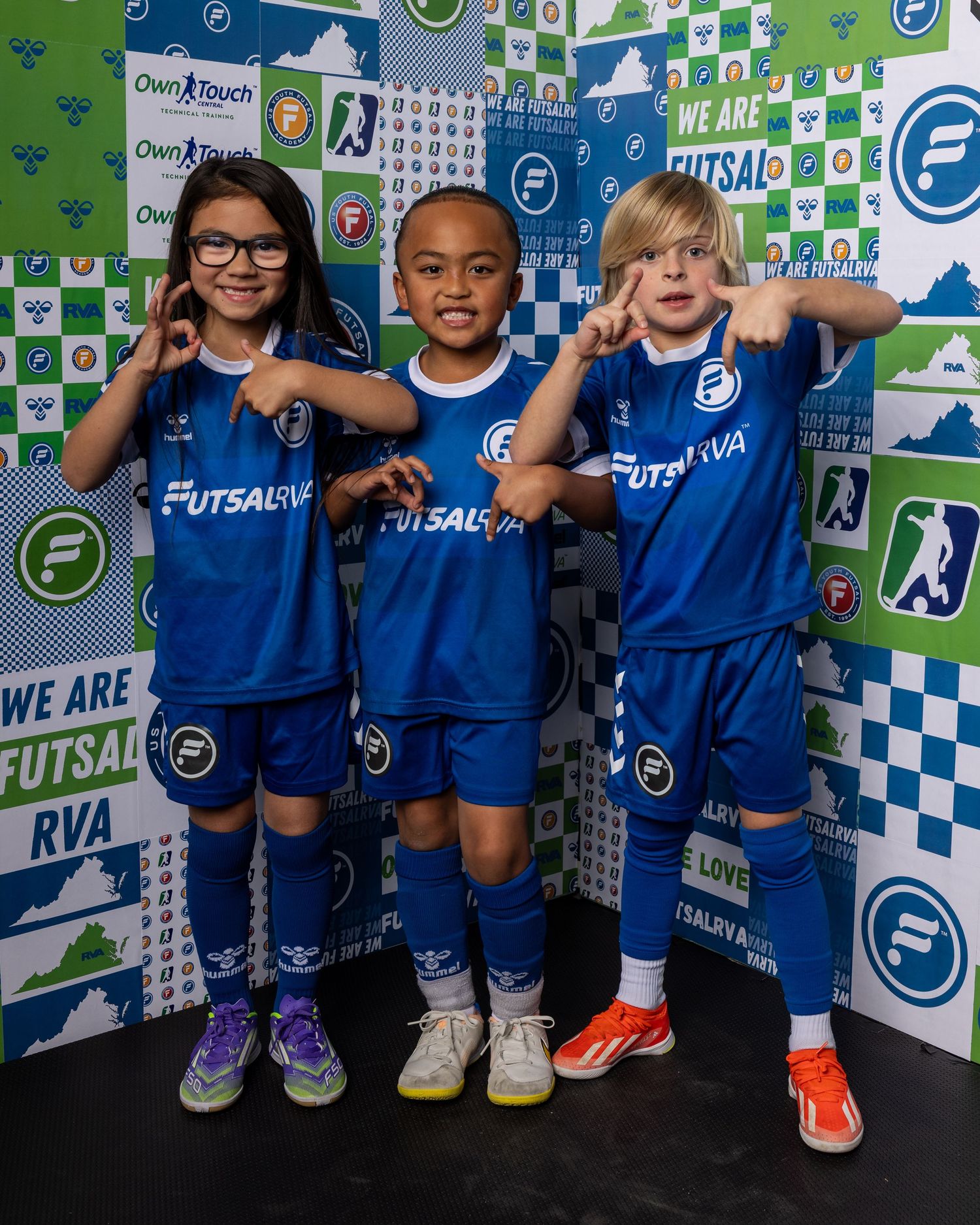 Young futsal players in blue uniforms pose together against a branded backdrop with team logos.