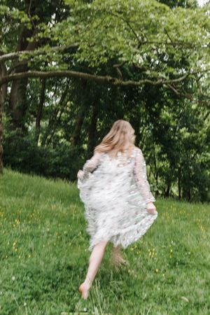A figure in a flowing white dress twirls playfully in a sunlit meadow surrounded by lush green trees.