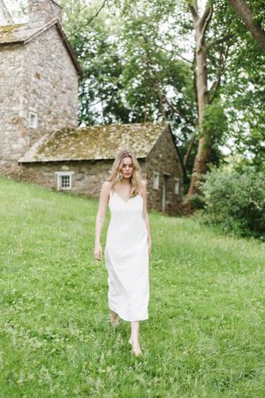A woman in a flowing white dress walks down a grassy hill near stone buildings on a sunny summer day.