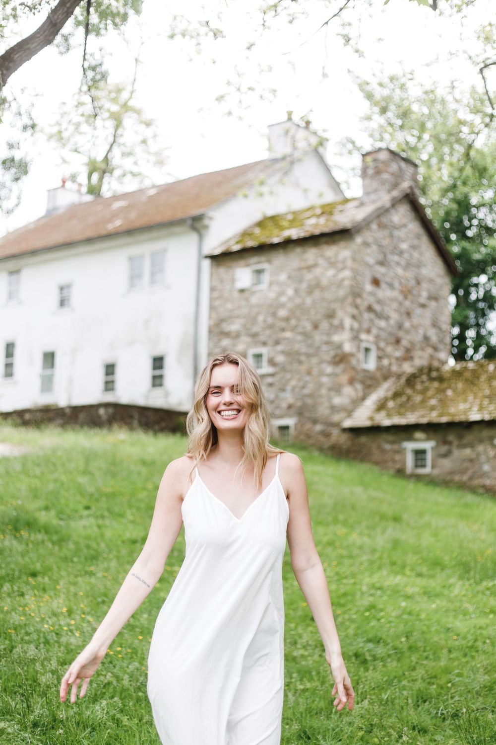 A carefree woman in a white sundress dances joyfully on a green hillside near rustic stone buildings.
