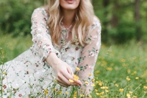 Someone wearing a delicate floral dress sits in a blooming meadow picking wildflowers on a sunny spring day.