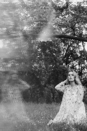 Artistic black and white portrait of a figure in a floral dress standing in a dreamy forest setting with soft light effects.