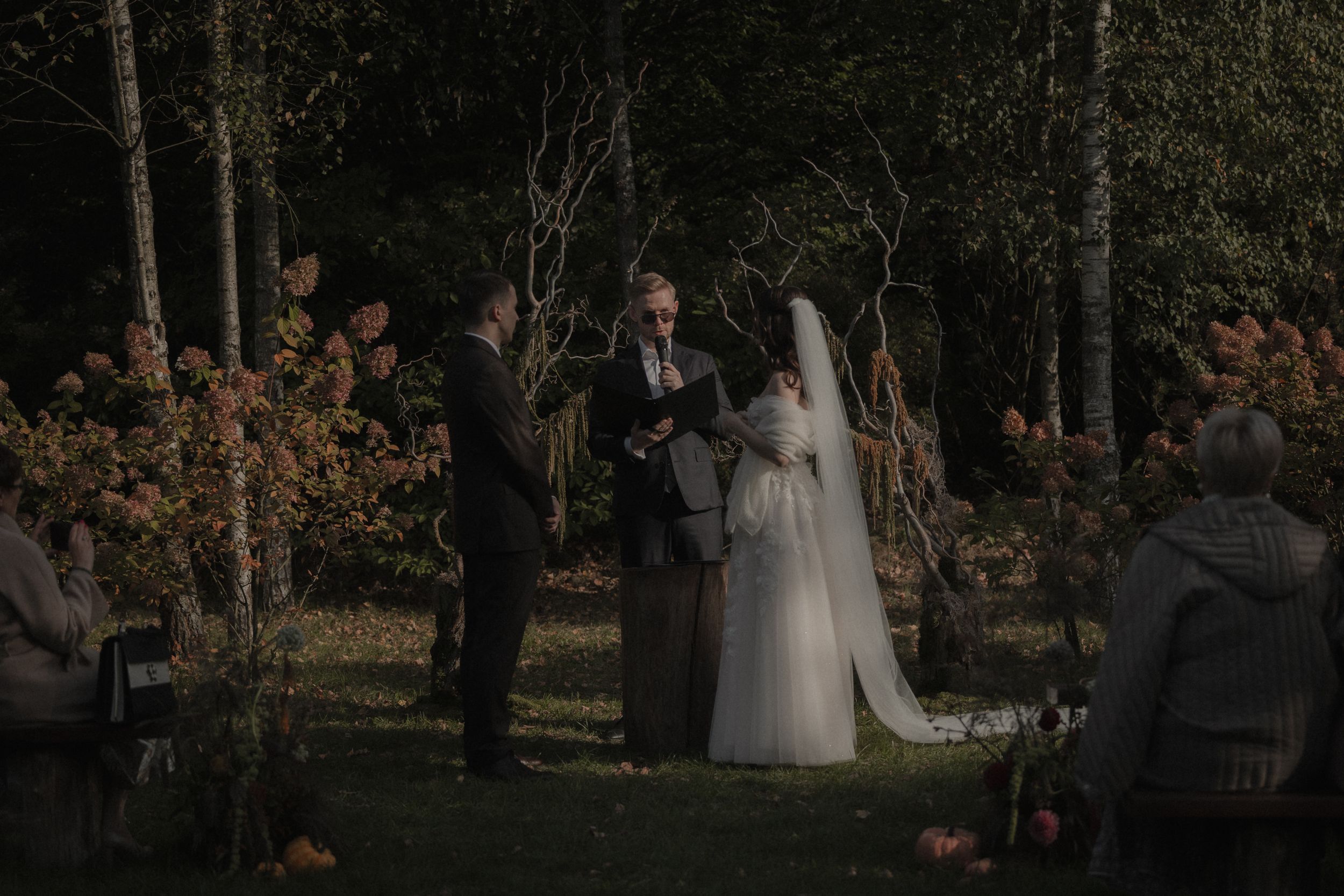 A couple in wedding attire walks through a wooded area with fall foliage during an outdoor ceremony.