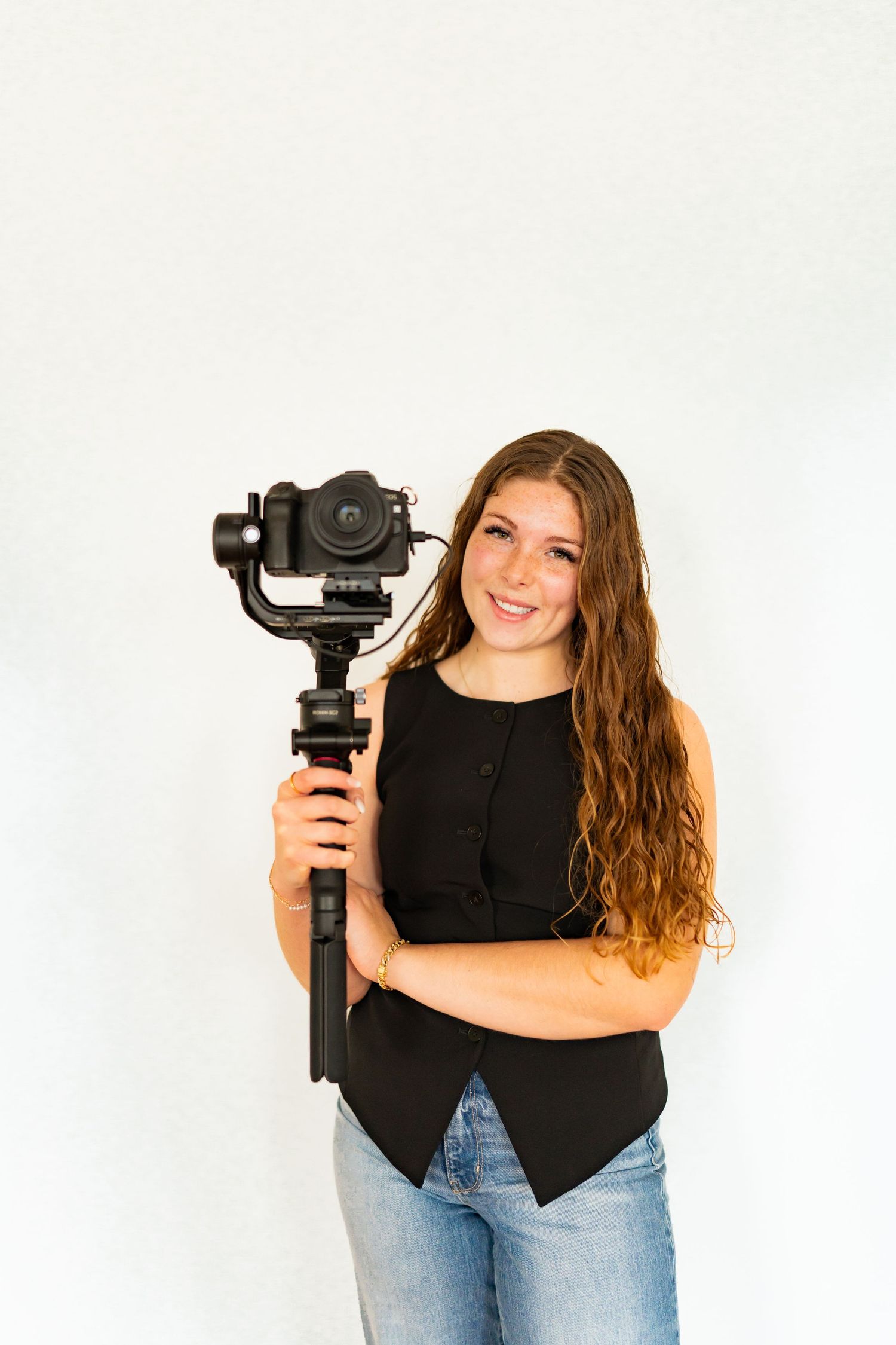 Camera operator in black top and jeans holds professional camera equipment against white background.
