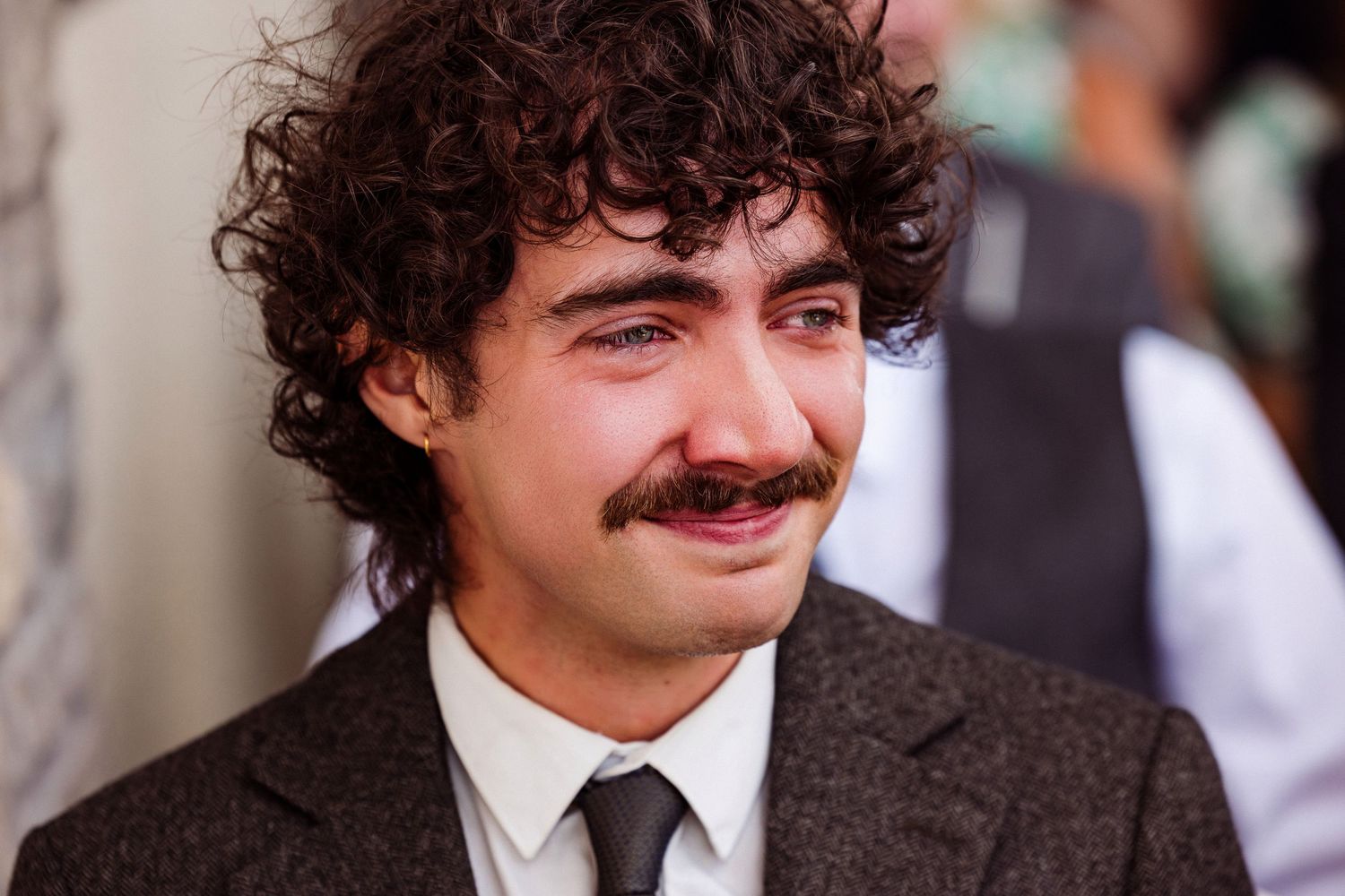 A young man with curly dark hair and a mustache wearing a brown suit and tie smiles at the camera.