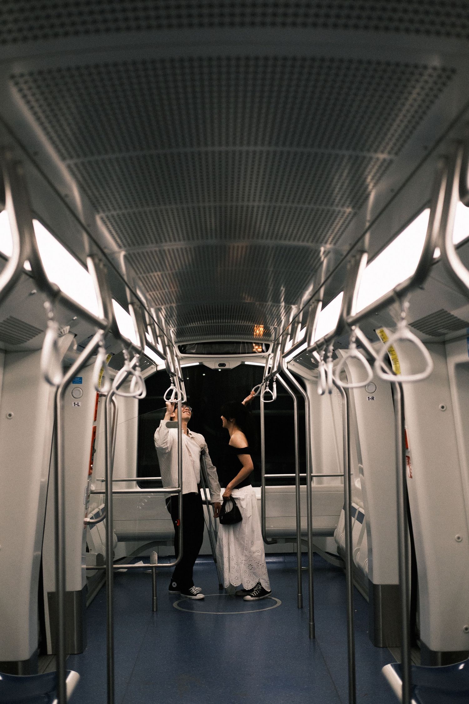 Interior view of a modern subway car with white walls, handrails, and dim lighting showing multiple passengers in motion.