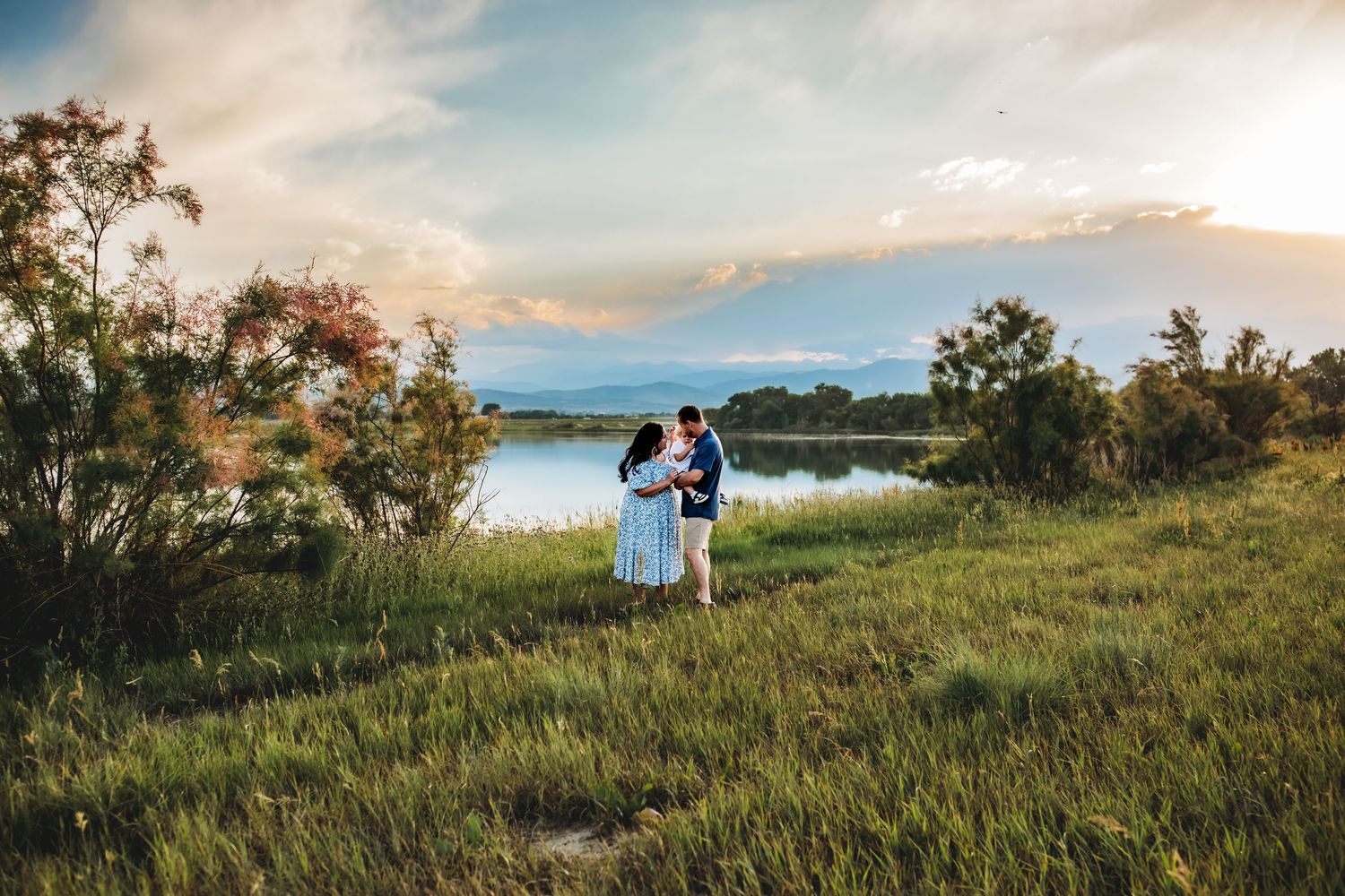 Two people stand together on a grassy hill overlooking a serene lake at sunset with trees in the background.