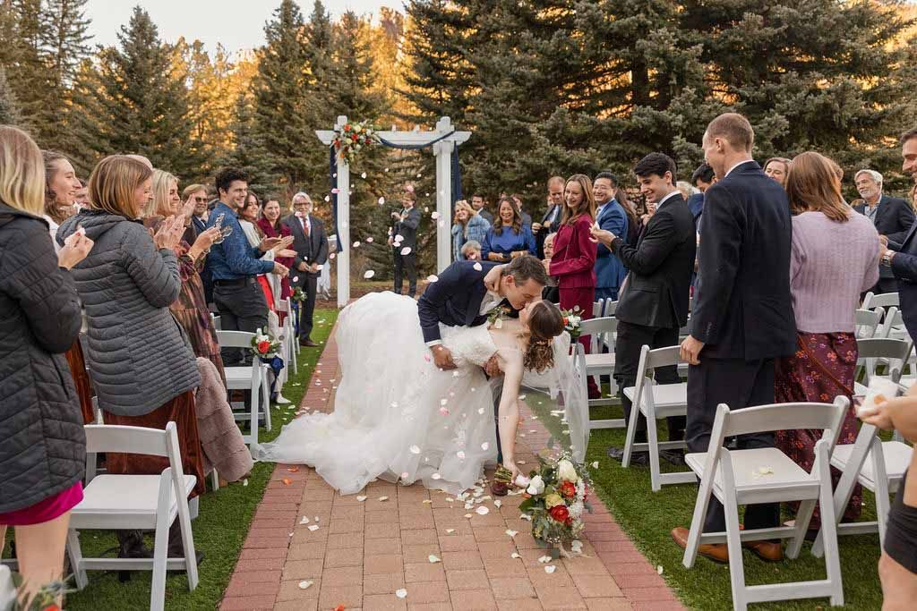 A dog walks down the aisle during an outdoor autumn wedding ceremony with white chairs and guests gathered.