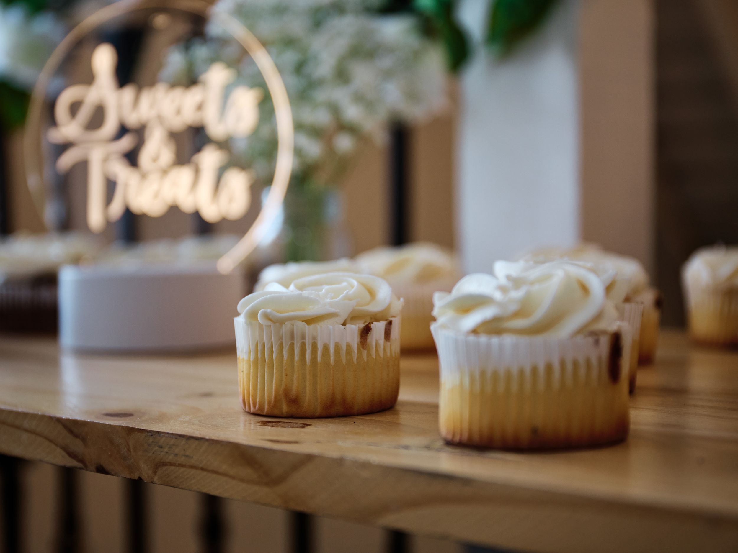Two vanilla cupcakes with swirled white frosting sit on a wooden surface with a neon sign glowing in the background.