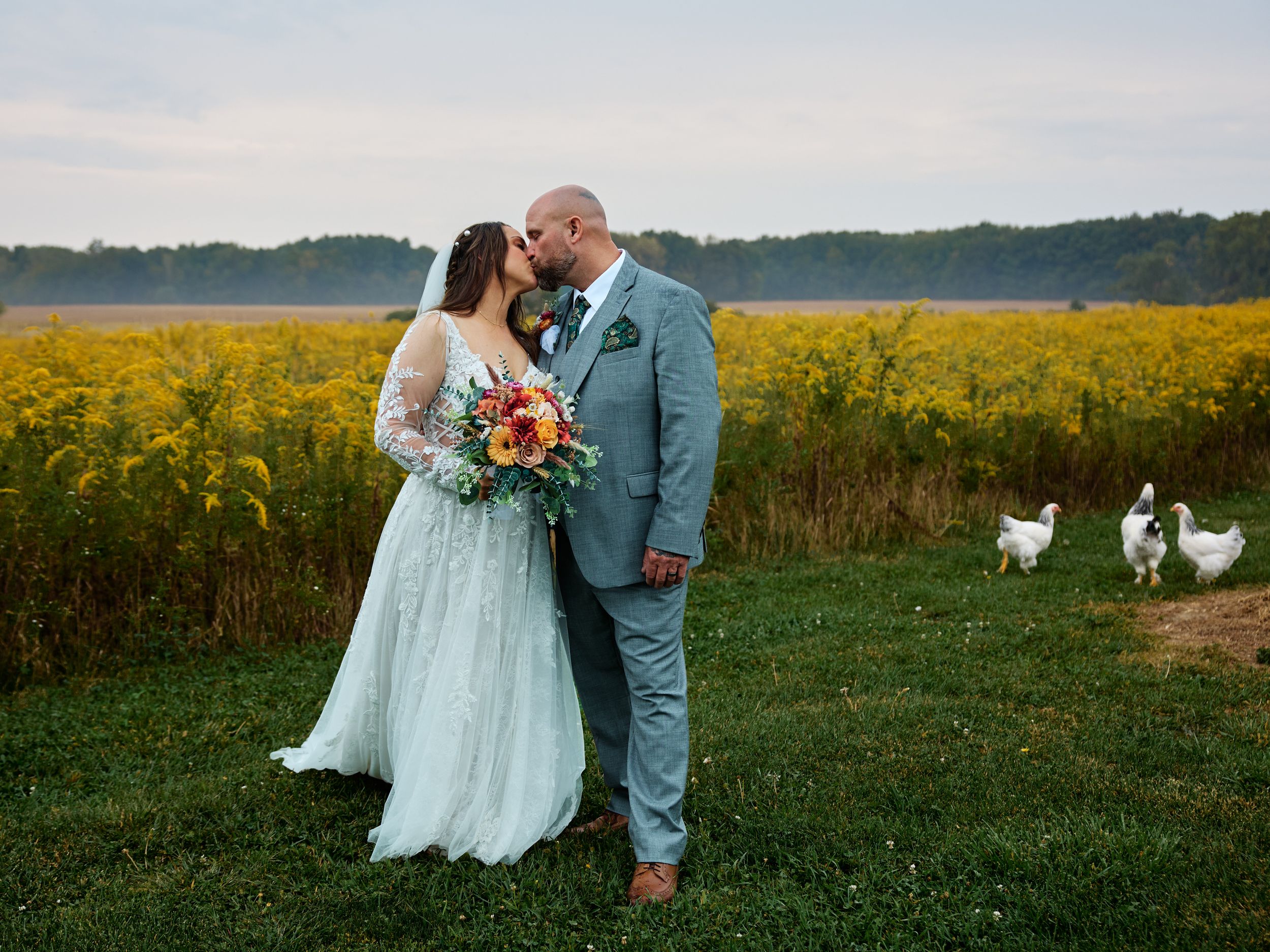 A romantic couple in a mint green dress and blue suit embrace in a field of golden sunflowers and rustic farmland.