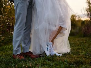Newlywed couple's feet shown together in sneakers and formal wedding attire on grassy field at sunset.