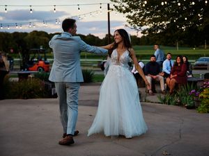 A couple in wedding attire dances together on an outdoor patio with string lights overhead during sunset.