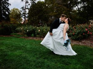 A couple in wedding attire dances and twirls together on lush green grass near blooming gardens.