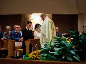 A couple sits in a church pew during a ceremony while a priest in green vestments stands nearby with decorative plants visible.