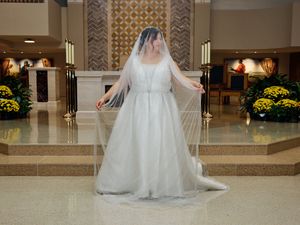 A person in a flowing white wedding dress and veil stands gracefully in an elegant lobby with yellow flowers and columns.
