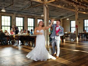 Two people dance together during a wedding reception in a rustic venue with wooden floors and string lights.