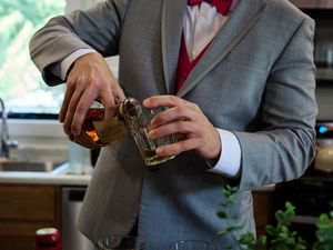 A person in a gray suit and red tie holds a glass of whiskey while sitting at a bar.
