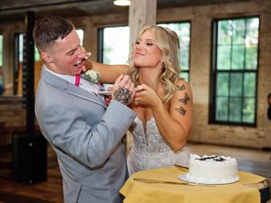 Newlywed couple feeds each other wedding cake during a reception in a rustic venue with exposed brick walls.