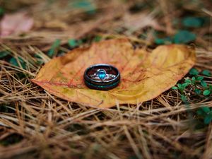 Wedding ring rests on a fallen autumn leaf against a blurred natural background of pine needles and grass.