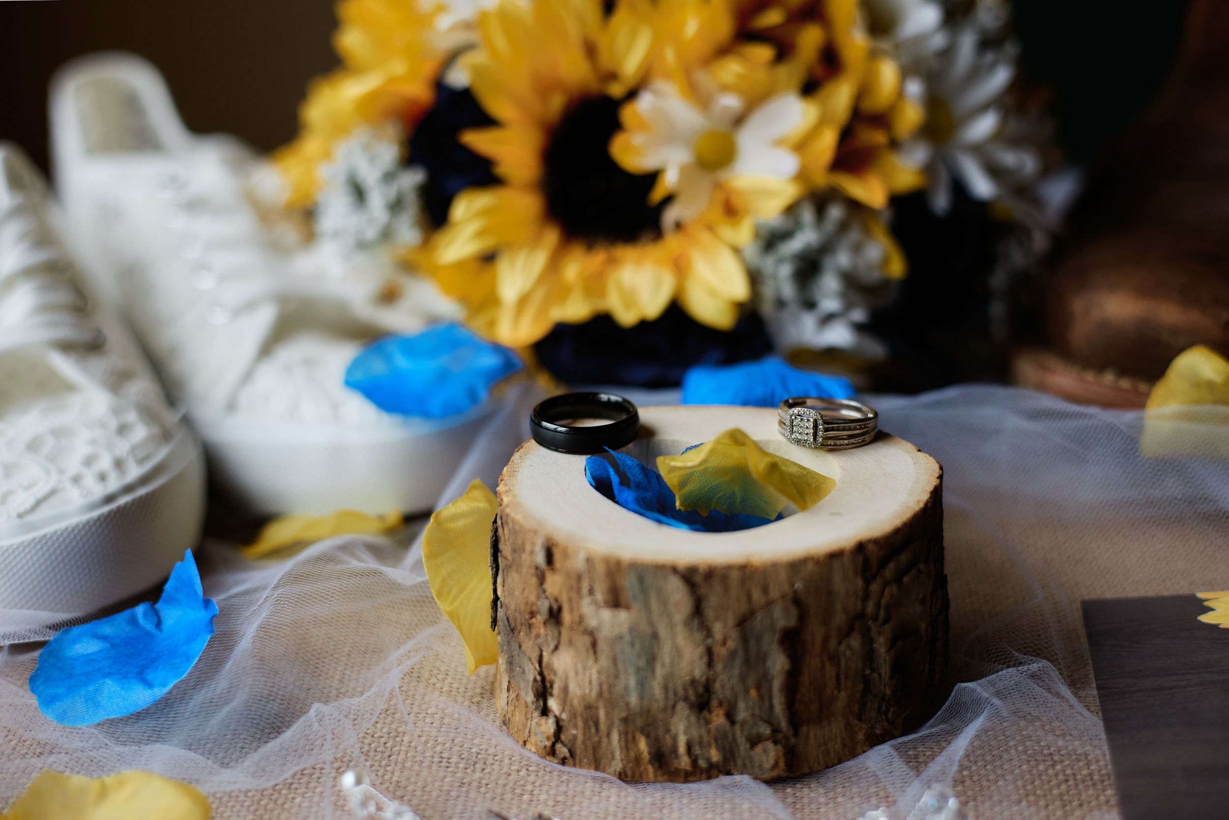 A rustic wedding ring display with sunflowers and blue butterflies on wooden slices atop white tulle fabric.