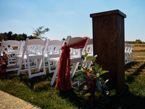 White wooden chairs and red floral arrangements line up at an outdoor wedding ceremony against a blue sky.